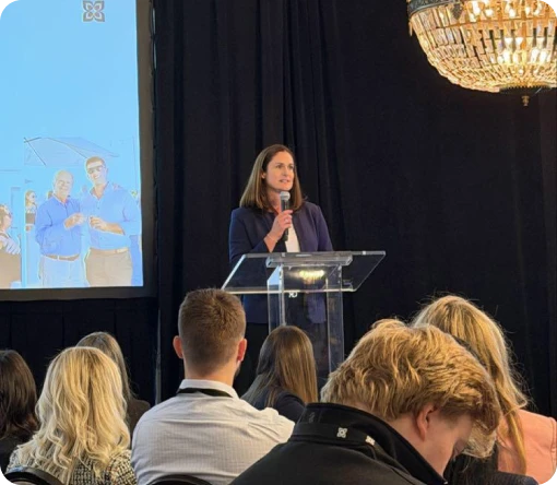 A speaker presents at a conference, with an audience engaged and a large screen displaying images in the background.