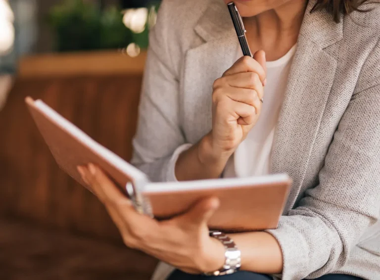 A woman in a blazer thoughtfully writes in a notebook while holding a pen, reflecting on her ideas.