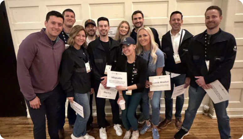 A diverse group of professionals poses together, holding signs with words like "Adaptable" and "Growth Mindset" at an event.