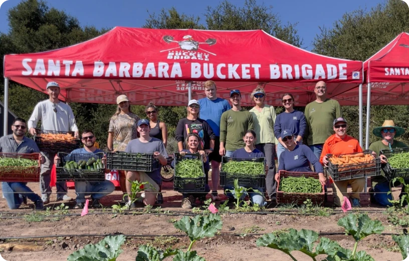 Volunteers from the Santa Barbara Bucket Brigade pose with harvested vegetables under a red canopy in a community garden.