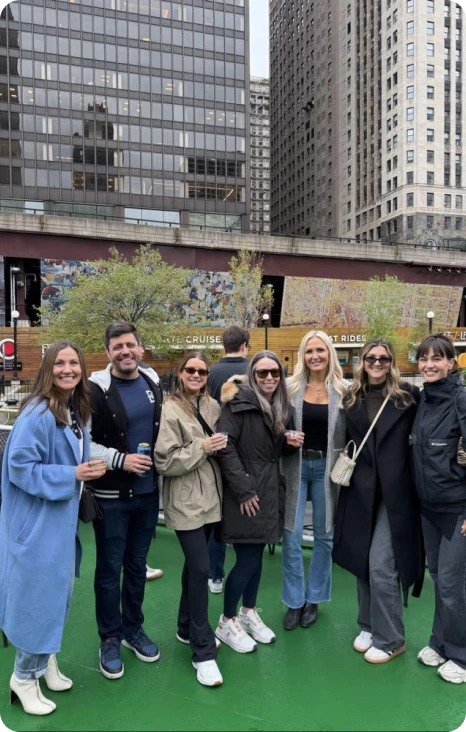 A group of seven friends posing together on a rooftop with drinks, surrounded by tall buildings and greenery.