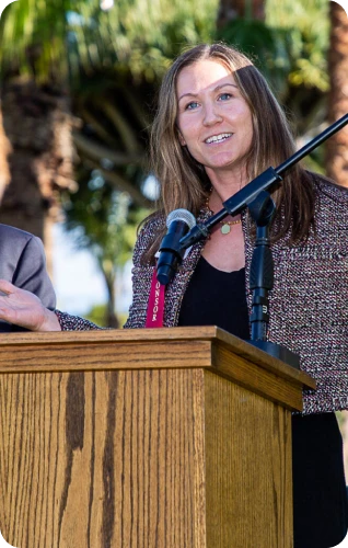 A woman speaks at a podium outdoors, engaging the audience with a smile and a microphone in hand.