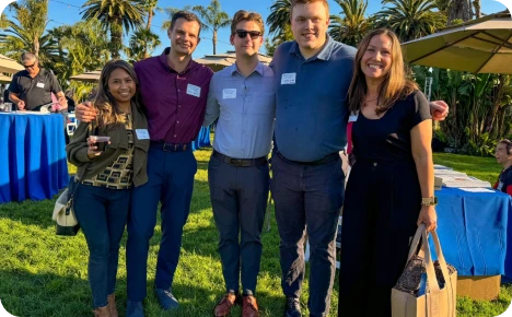 Five professionals pose together at an outdoor networking event with palm trees and tables in the background.