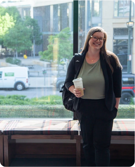 A smiling woman in a blazer holds a coffee cup while standing by a large window with a city view.