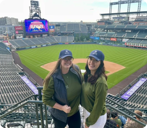 Two women in matching outfits and hats pose together at a baseball stadium with a large screen and green field in the background.