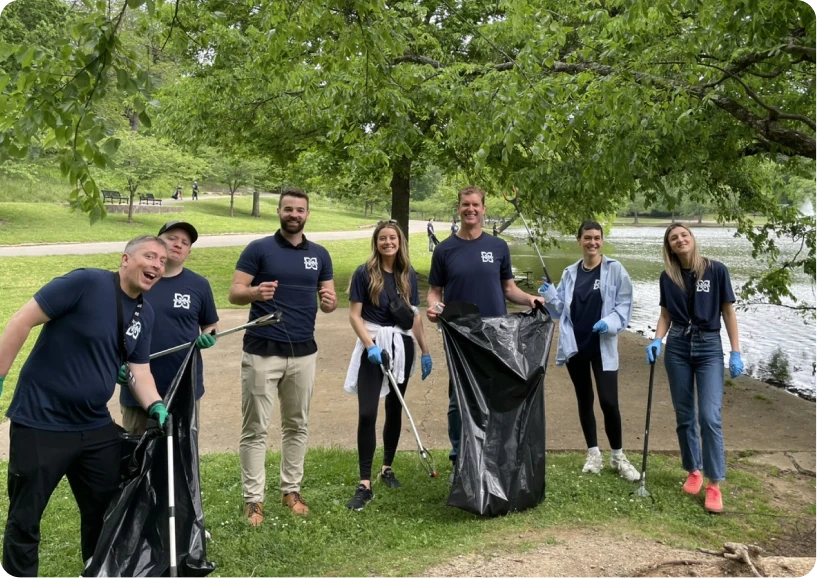 Volunteers in matching shirts gather by a lake, holding trash bags and tools for a community cleanup event.