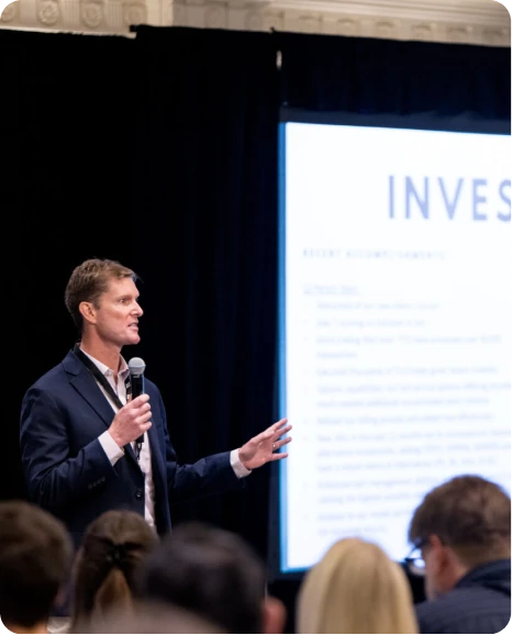 Presenter speaking to an audience with a slide titled "Investments" displayed in the background.