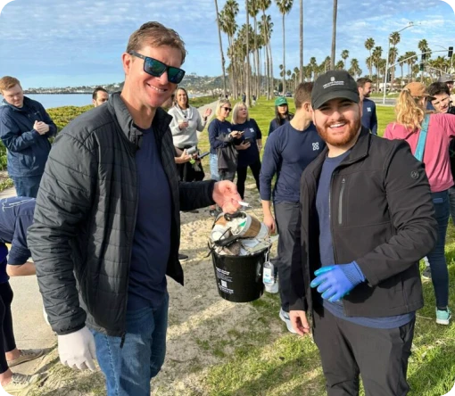 Two volunteers smile while holding a bucket of collected trash during a beach cleanup event, with palm trees in the background.