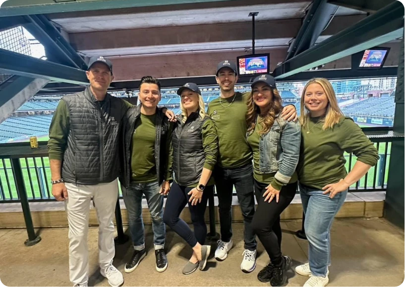 A group of six friends poses in matching green outfits and hats at a sports stadium, ready for a game day celebration.