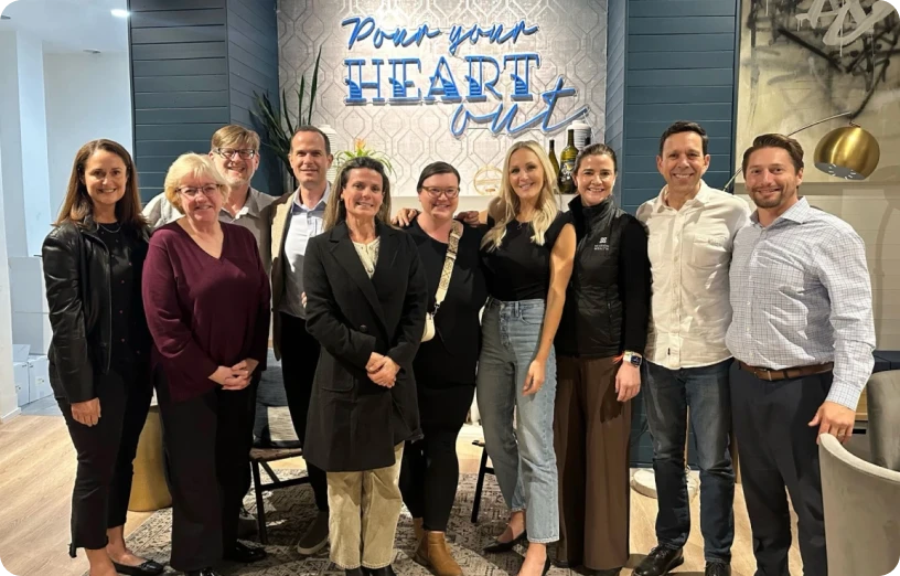 A diverse group of professionals poses together in a welcoming space with a "Pour your HEART out" sign in the background.