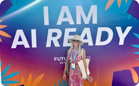 A woman in a floral dress and sunhat stands beside a colorful backdrop that reads "I AM AI READY."