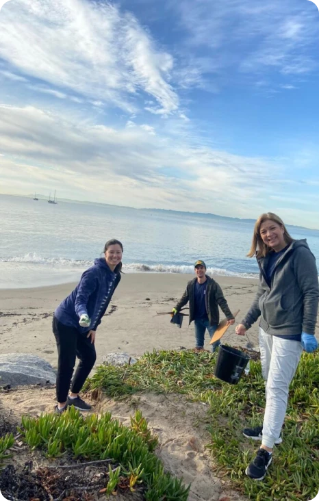 Three volunteers clean the beach, holding tools and smiling, with a calm ocean and cloudy sky in the background.