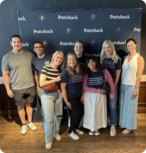 A diverse group of eight people smiles together in front of a Puttshack backdrop, showcasing a fun social gathering.