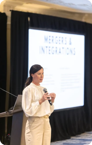 A speaker presents on mergers and integrations at a conference, with a presentation displayed in the background.