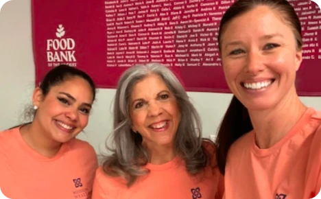 Three smiling women in orange shirts pose together in front of a Food Bank poster, promoting community service and support.