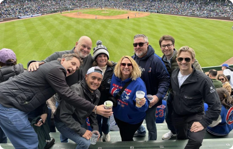 Group of friends enjoying a baseball game, smiling and holding drinks in a crowded stadium with the field visible.