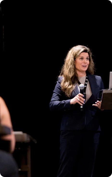 A woman in a suit speaks confidently at a podium during a presentation, engaging with her audience.