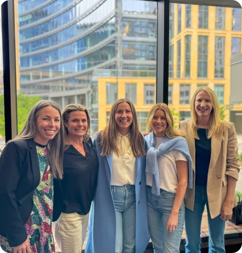 A group of five women stands together, smiling, against a backdrop of modern buildings and large windows.