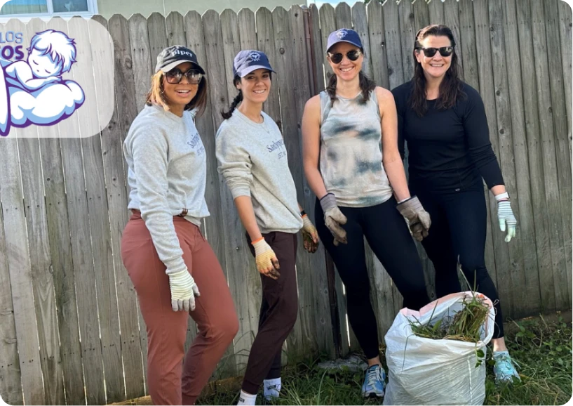 Four women in work attire smile while holding gardening gloves and a bag of weeds, standing in a backyard.