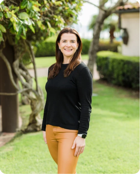 Smiling woman in a black long-sleeve shirt and orange pants stands outdoors among greenery, exuding confidence and approachability.