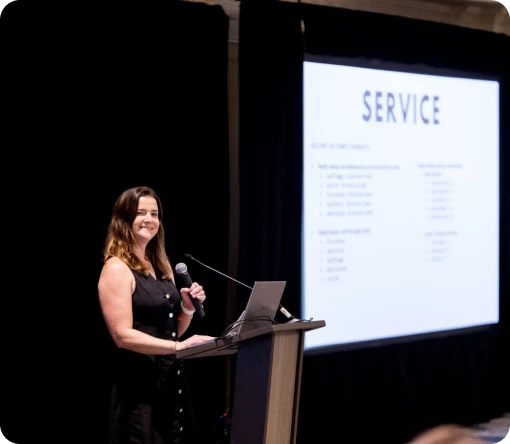 A woman presents at a podium with a presentation slide titled "SERVICE" visible in the background.