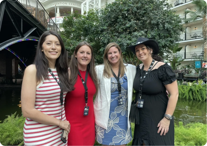 Four women pose together in a lush indoor garden near a water feature, all wearing event badges.