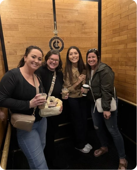 Four friends enjoying drinks and snacks together in a wooden elevator, smiling and posing for the camera.