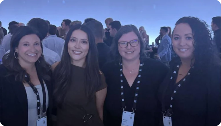 Four women pose together at a networking event, smiling against a backdrop of a large crowd and blue lighting.
