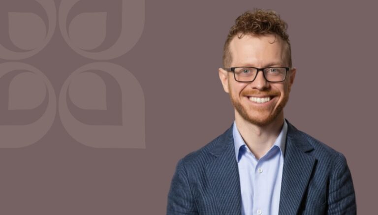 Smiling man in a blazer with glasses against a muted background, conveying professionalism and approachability.