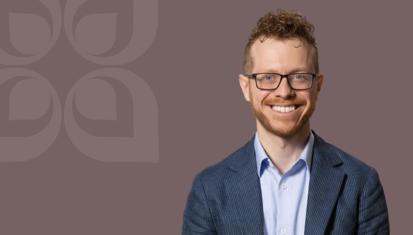 Smiling man in a blazer with glasses against a muted background, conveying professionalism and approachability.
