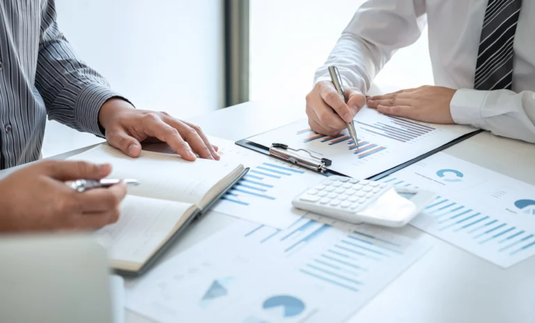 Two professionals analyze financial graphs and data during a market update meeting, with notes and a calculator on the table.