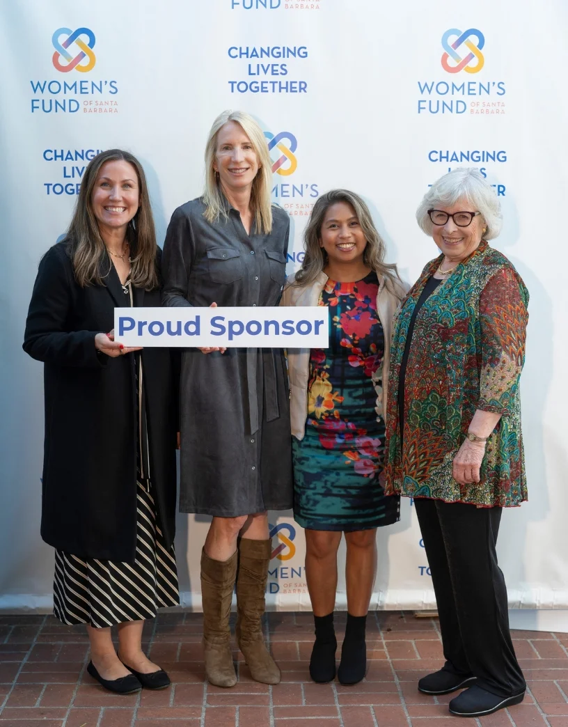 Four women pose together holding a "Proud Sponsor" sign in front of a Women's Fund backdrop at a community event.