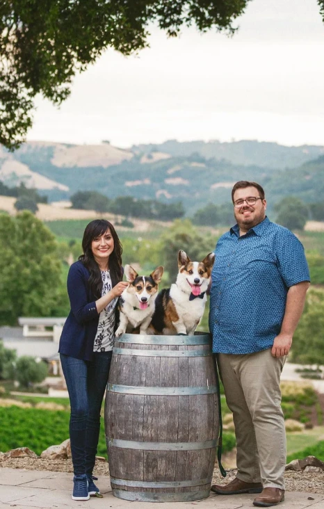 A couple poses with two corgis on a wooden barrel, set against a scenic vineyard landscape.