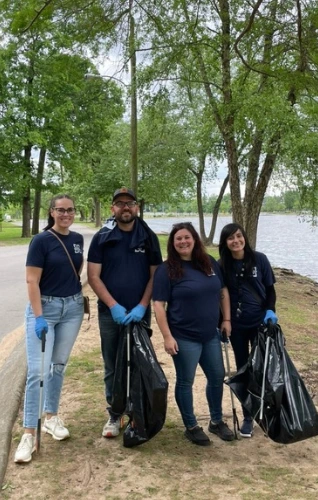 Four volunteers in matching shirts collect trash by a river, promoting community clean-up and environmental stewardship.