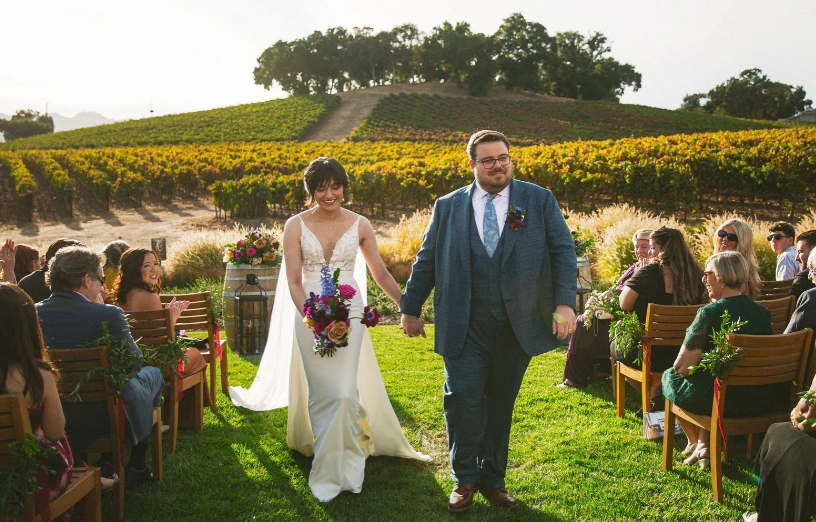 A joyful couple walks hand-in-hand down the aisle, surrounded by a vineyard and seated guests celebrating their wedding.