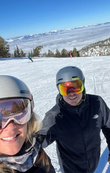 Two skiers smile for a selfie on a snowy slope with mountains and clouds in the background, enjoying a sunny day.