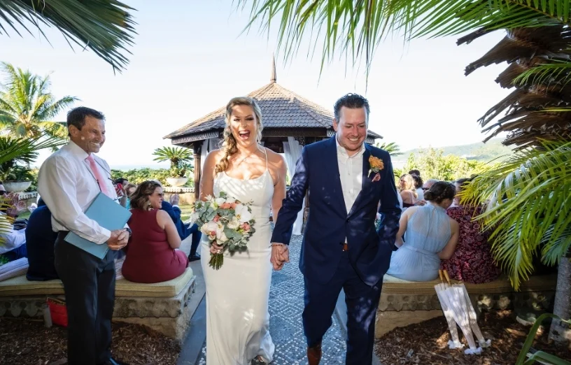 Joyful newlyweds walk hand-in-hand down a tropical aisle, surrounded by guests in a picturesque outdoor setting.