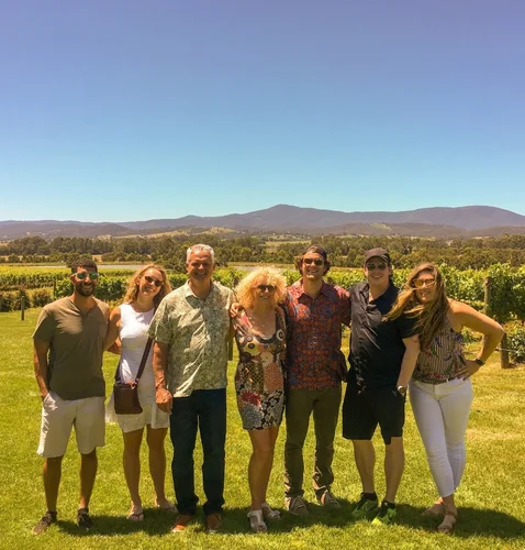 A group of seven friends poses together in a vineyard, with mountains and clear blue skies in the background.