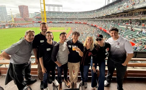 A group of eight people smiling at a baseball game, with a packed stadium and field in the background.