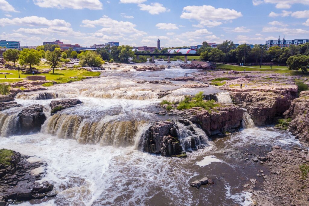 Waterfalls cascade over rocky formations in Sioux Falls, with lush greenery and city buildings in the background.