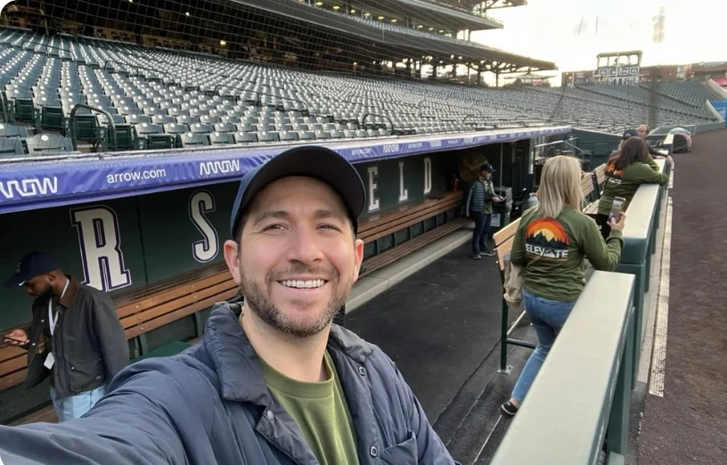 A man smiles for a selfie in a nearly empty stadium, with others socializing in the background.