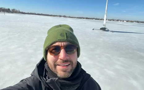 Smiling man in a green beanie stands on a frozen lake with a sailboat in the background under a clear blue sky.