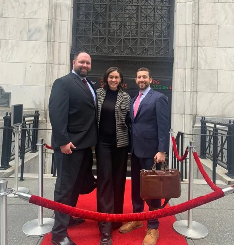 Three professionals pose on a red carpet outside a building, dressed in business attire, with a decorative entrance in the background.