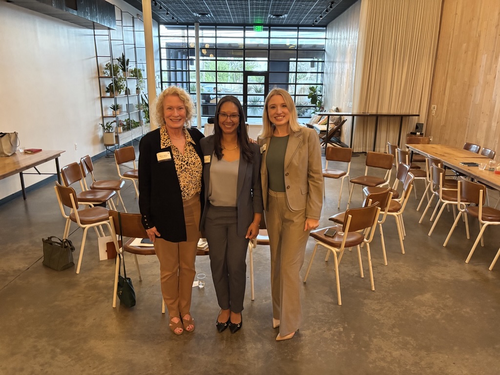 Three women stand together in a modern event space, promoting a discussion on women and wealth and financial well-being.